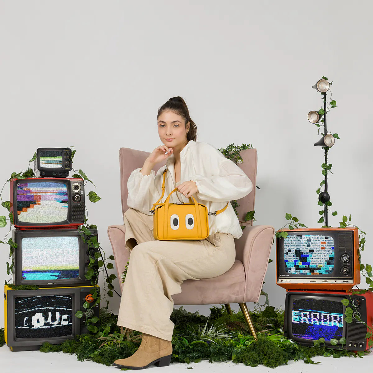 Model sitting in an armchair showcasing the Min and Mon Benji Golden handbag in yellow leather with large cartoon eyes. Retro set featuring vintage televisions and plants against a light background. Best Sellers banner.