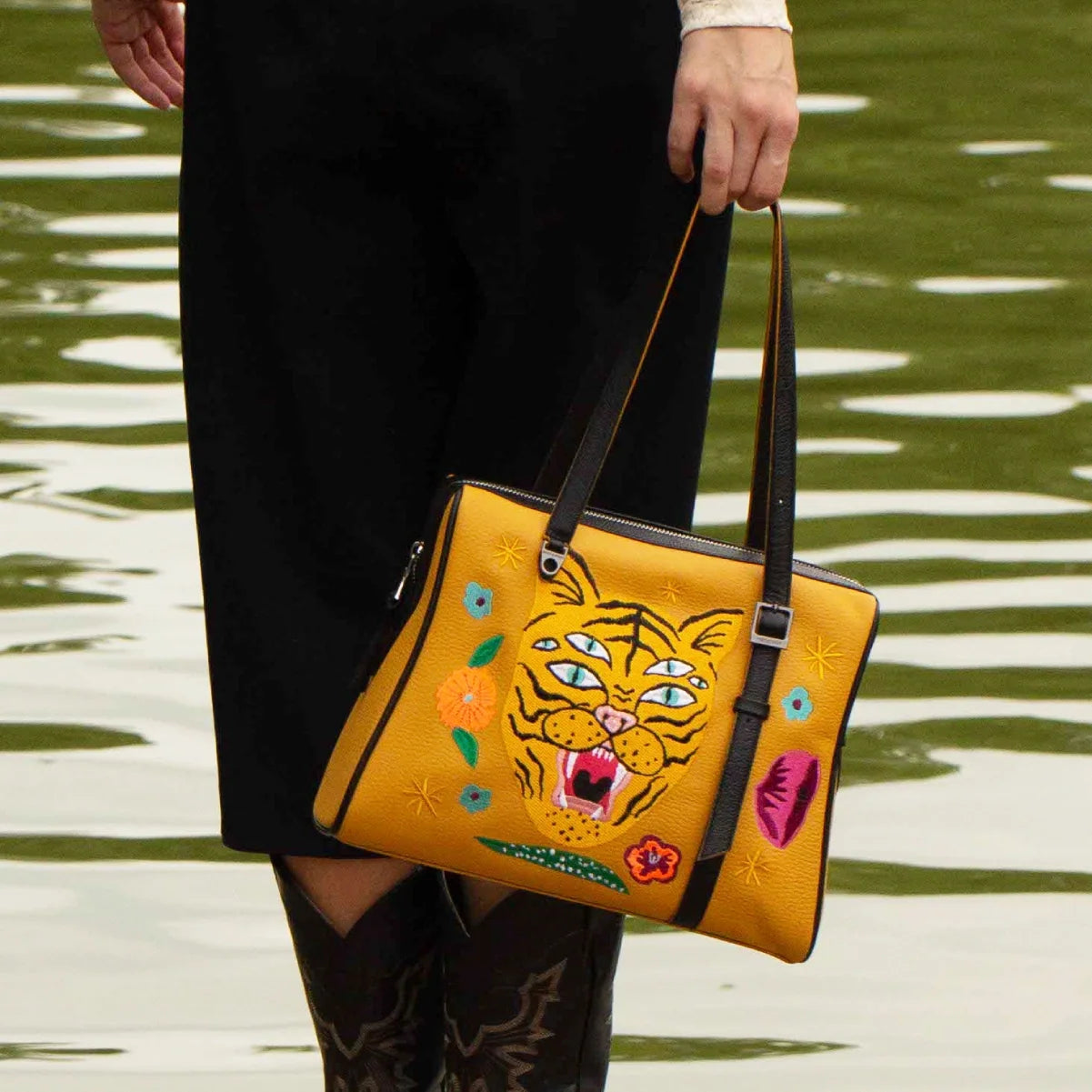 Close-up of the Min & Mon Esther Golden tote bag with a multicolored embroidered tiger, floral accents, and black shoulder straps, styled with a black outfit.