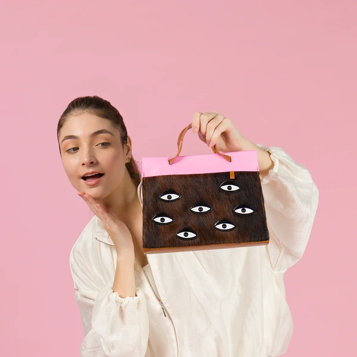 Surprised model holding a Min and Mon handbag from the All Over Eyes collection, featuring a brown textured cowhide body, embroidered eye patches, and a bright pink flap. Pink background.