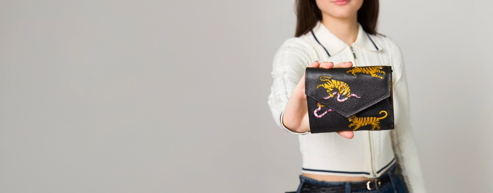 A woman in a black top and jeans holds a "Fiona Wallet" from the "El Trópico Collection" by Min & Mon, a NYC-based brand. The pink wallet features vibrant tiger head prints, highlighting its bold and unique design.