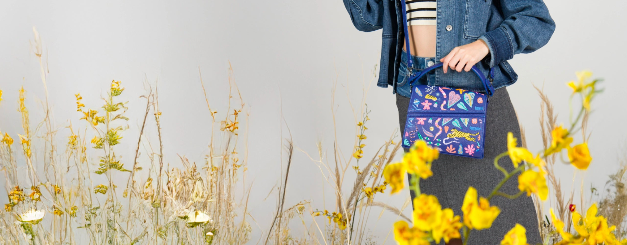 A woman in a white crochet top stands in a lush green forest, holding an "Anastasio Mini crossbody handbag" from the "El Trópico Collection" by Min & Mon, a NYC-based brand. The bright green handbag features vibrant, embroidered tropical designs.