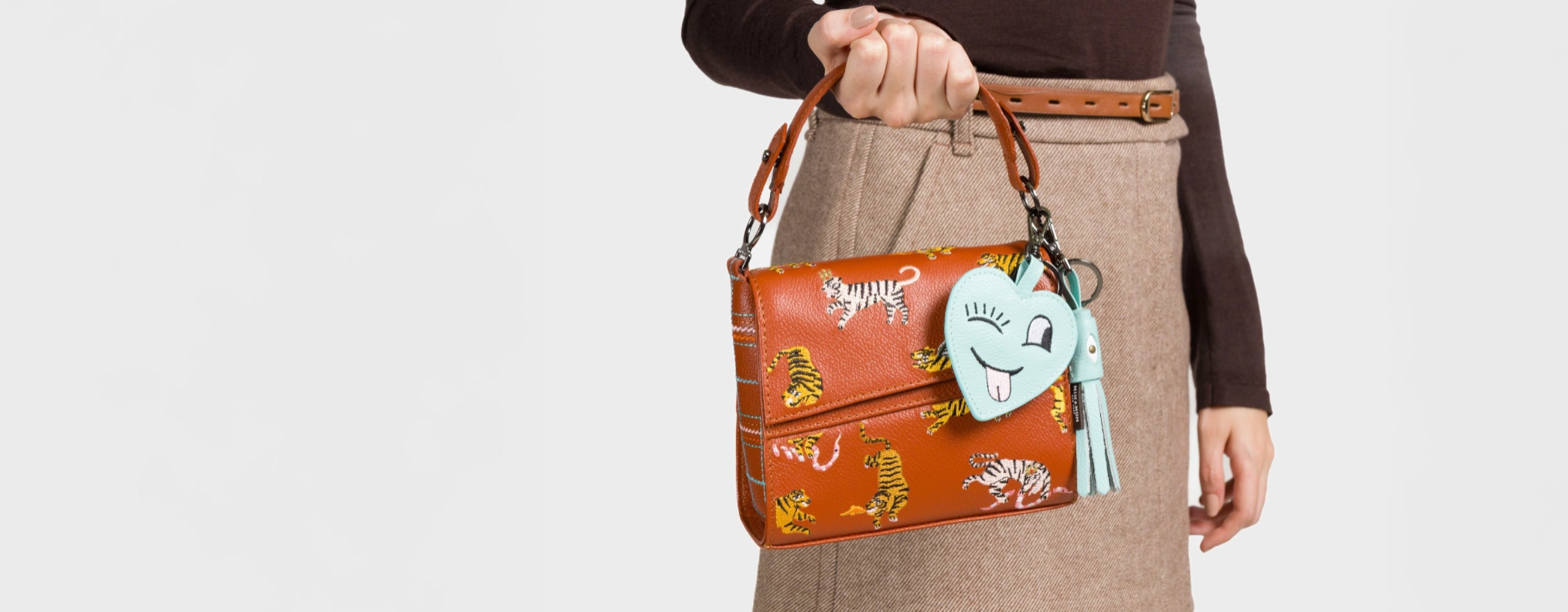 A woman with long brown hair wears a cream-colored knit sweater and a light blue denim skirt, holding a black "Vali Crossbody-handbag" by Min & Mon, a NYC-based brand. The handbag features a pink strap with eye designs and a pink tassel charm accessory.