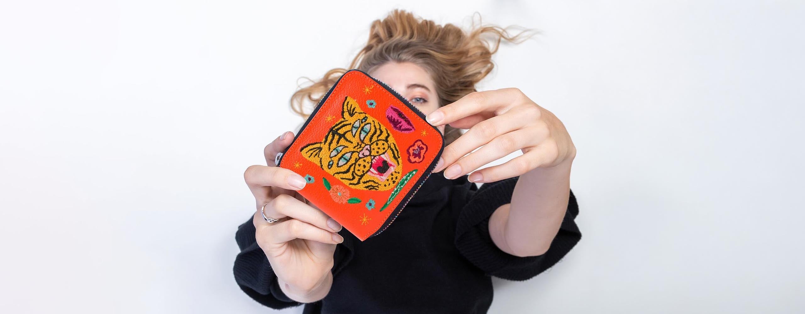 A woman in a black top and jeans holds a "Fiona Wallet" from the "El Trópico Collection" by Min & Mon, a NYC-based brand. The pink wallet features vibrant tiger head prints, highlighting its bold and unique design.