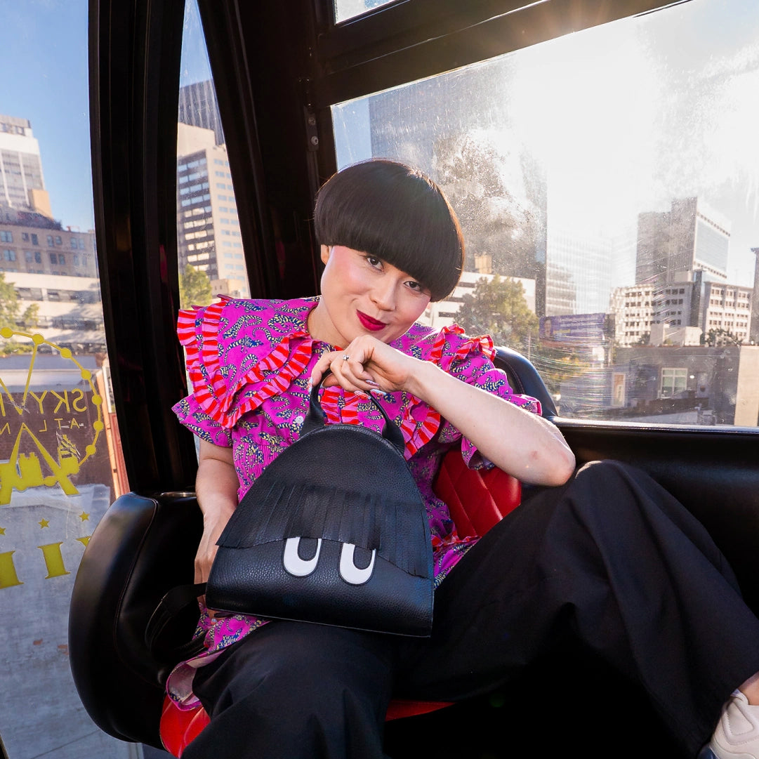 Comedian Atsuko Okatsuka sitting inside a New York City tram, wearing a vibrant pink patterned top and holding a black Min & Mon handbag featuring playful fringe and signature white eyes.