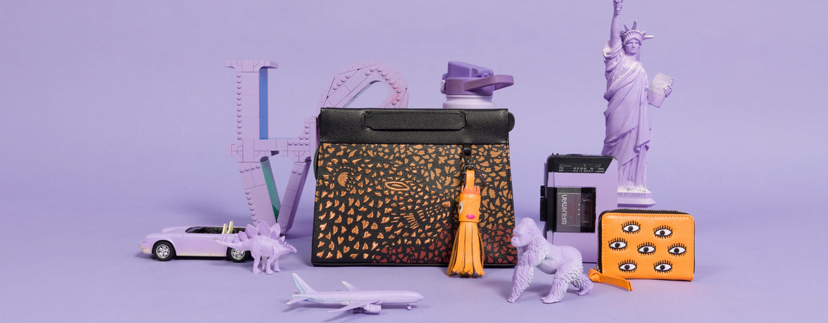 Min & Mon Vali handbag featuring a black and orange patterned design with a playful orange tassel charm, displayed alongside a matching orange Frodo wallet with embroidered eyes and whimsical purple accessories.
