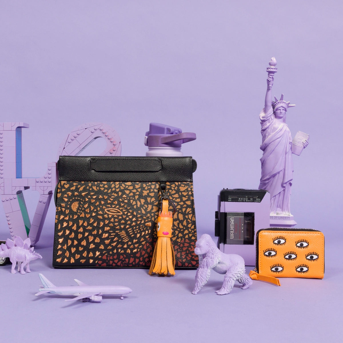 Close-up of a Min & Mon Vali handbag with a black and orange textured print, personalized with an orange tassel charm, next to an orange Frodo wallet featuring signature embroidered eyes, surrounded by playful purple objects.