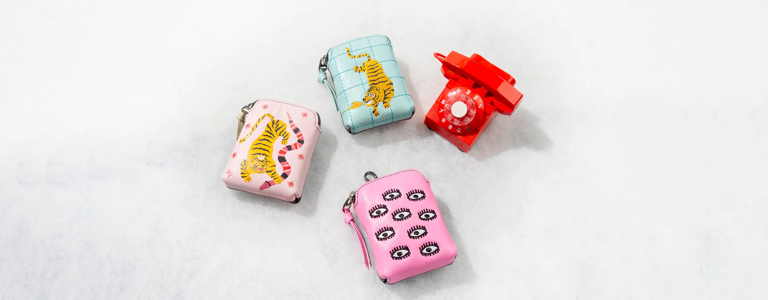 Small leather accessories with bold embroidered prints, arranged next to a red vintage rotary phone on a light background.