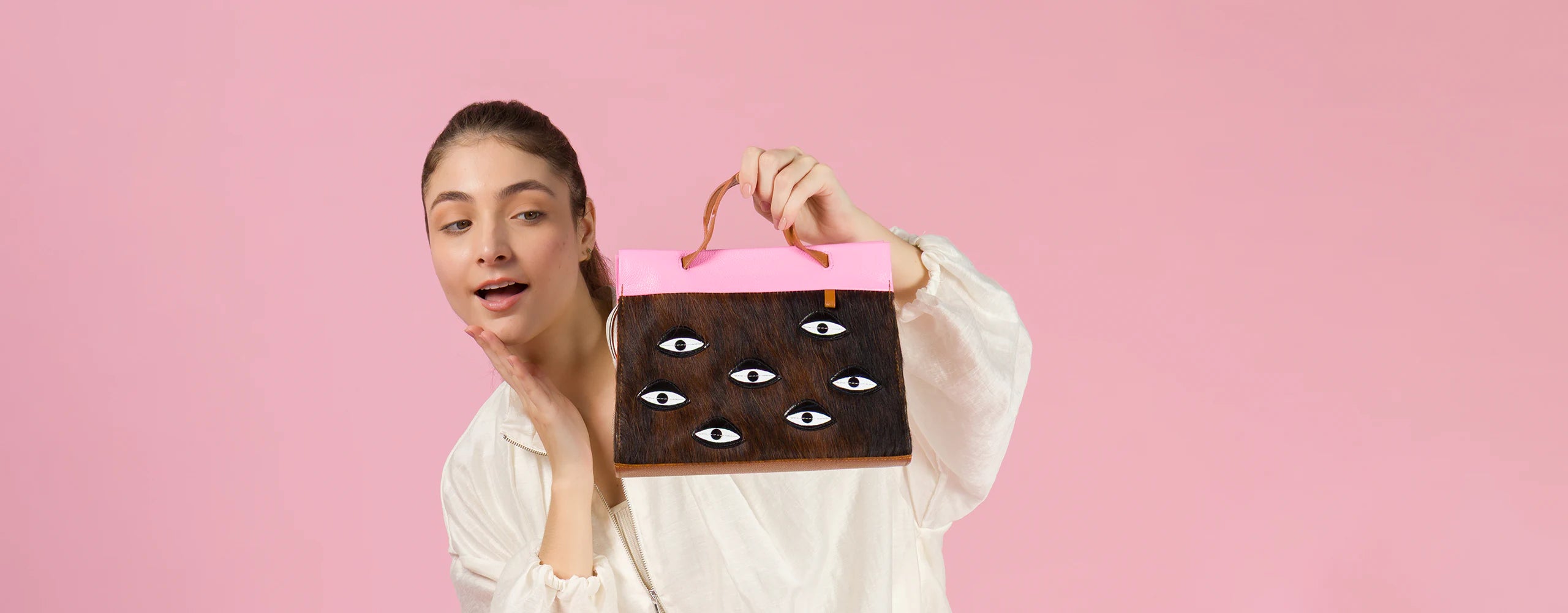 Surprised model holding a Min and Mon handbag from the All Over Eyes collection, featuring a brown textured cowhide body, embroidered eye patches, and a bright pink flap. Pink background.