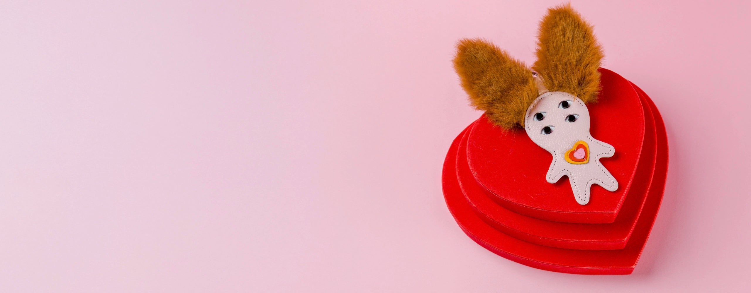 Red heart-shaped box with a whimsical Min & Mon leather charm featuring furry ears and a tiny heart detail on a pink background.