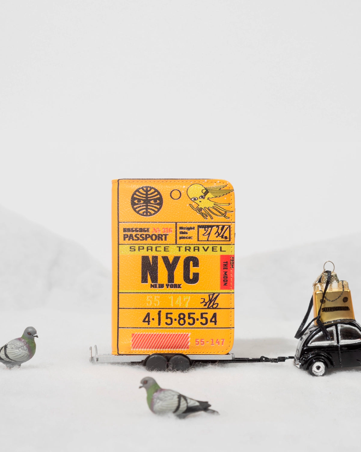 Yellow NYC passport-inspired wallet on a toy cart, with pigeon figurines around it on a white winter background.