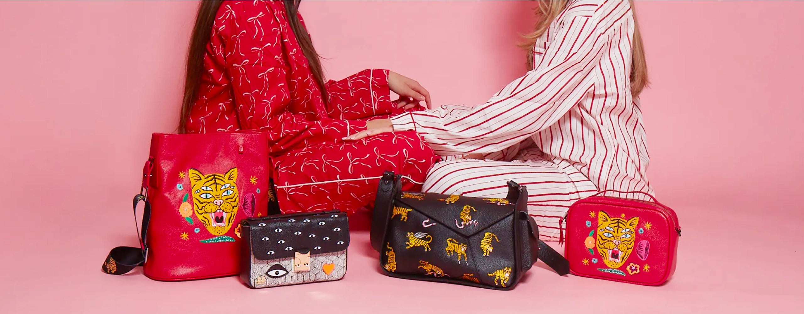 Two women sitting face to face in red and white pajamas, surrounded by Min & Mon bags and accessories with tiger and eye embroidery on a pink background.