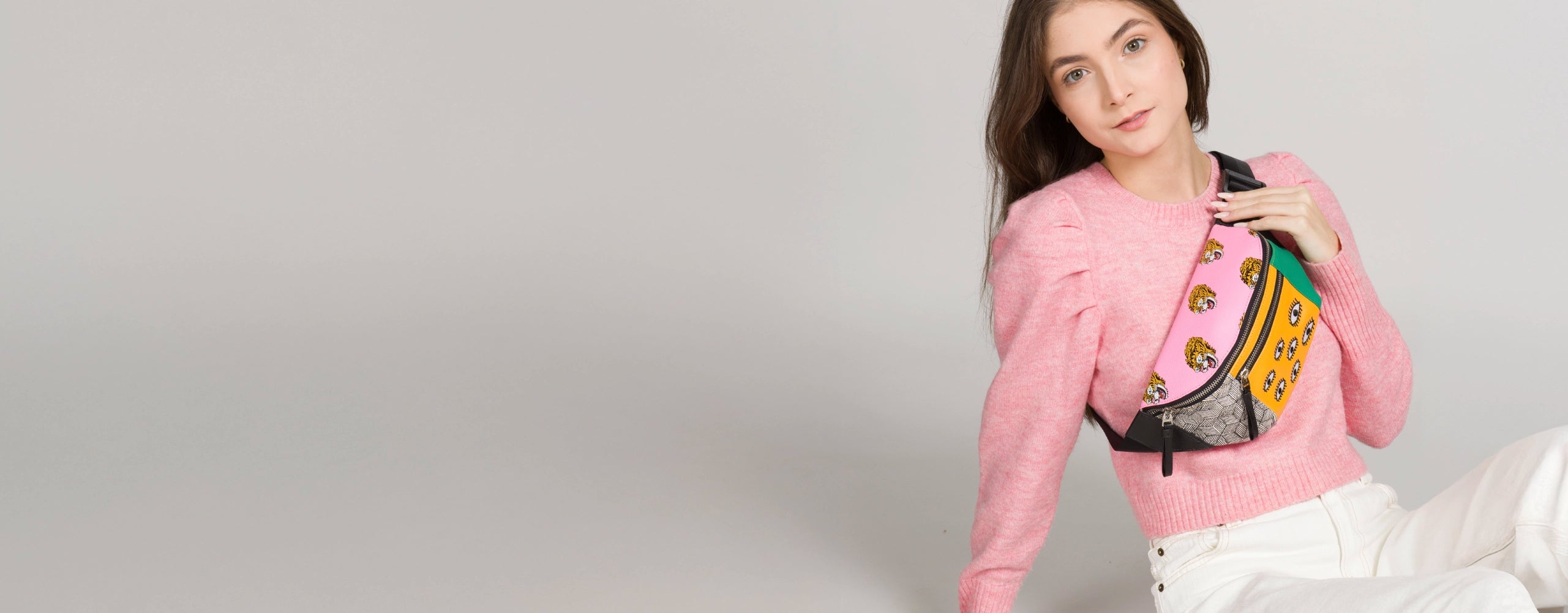 Model wearing a pink sweater and white pants, styled with a colorful Min & Mon crossbody bag featuring tiger and eye prints on a neutral background.
