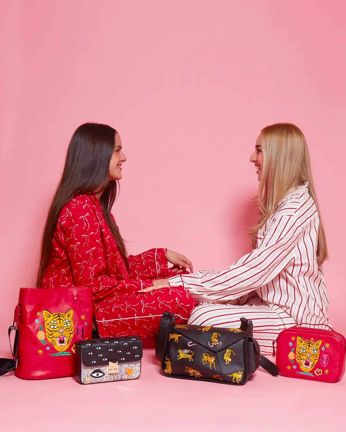 Two women smiling at each other while sitting on the floor with embroidered red and black Min & Mon bags, creating a warm and festive scene on a pink background.