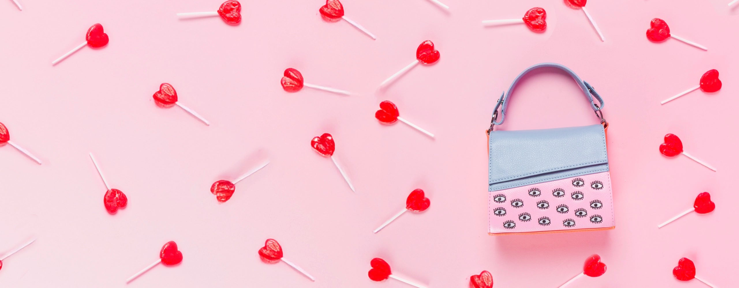 Mini shoulder bag in pastel blue and pink with embroidered eyes, styled on a pink background with red heart-shaped lollipops scattered around.