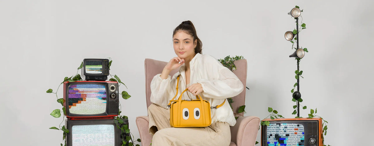 Model sitting in an armchair showcasing the Min and Mon Benji Golden handbag in yellow leather with large cartoon eyes. Retro set featuring vintage televisions and plants against a light background. Best Sellers banner.