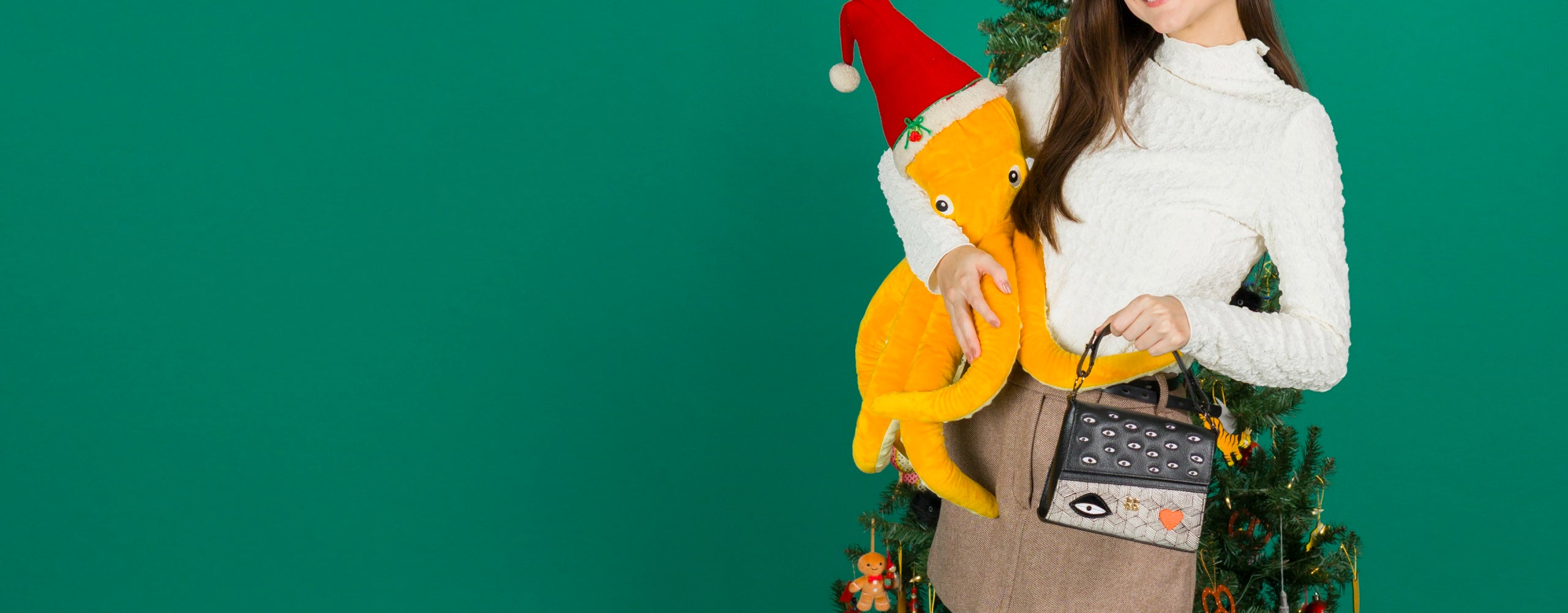 Model holding a Min & Mon crossbody bag with eye motif, standing by a decorated Christmas tree and holding a yellow octopus plush wearing a Santa hat against a green background.