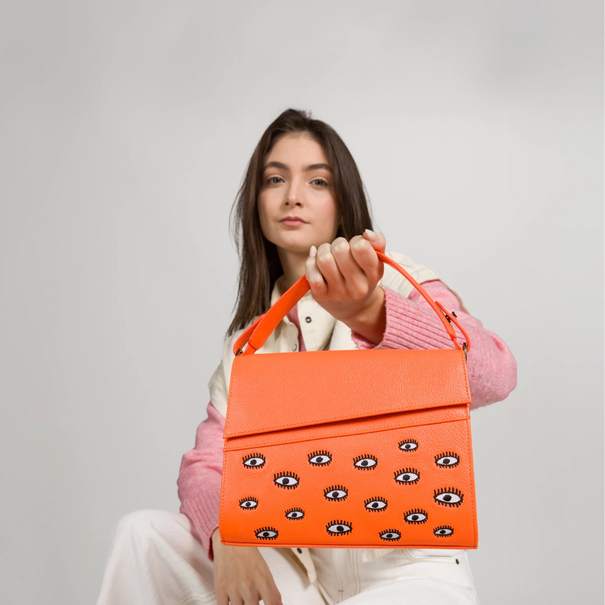 Seated model showcasing an orange bag with embroidered eyes, casual styling with pink sweater and light pants.