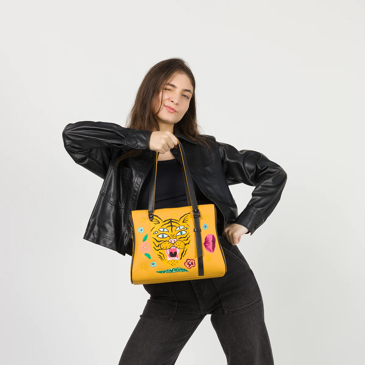 Model winking in a black leather jacket, holding the Min and Mon Esther Happy Tiger handbag in yellow leather featuring a colorful embroidered tiger from the Creatures of the Future collection. Light gray background.