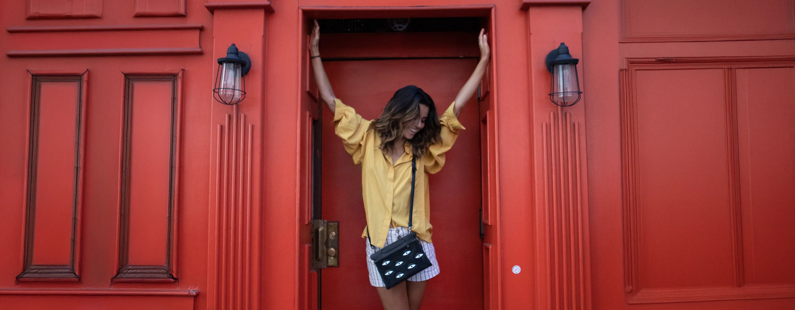 Woman posing in front of a bold red doorway wearing a black eye-detail crossbody bag.