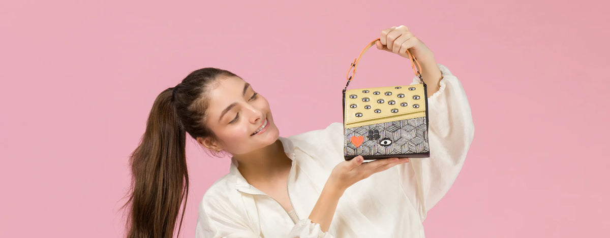 A smiling model in a white blouse holds a Min and Mon handbag featuring a yellow textured flap with eye motifs, a black-and-white geometric pattern, and a red heart patch. Pastel pink background.