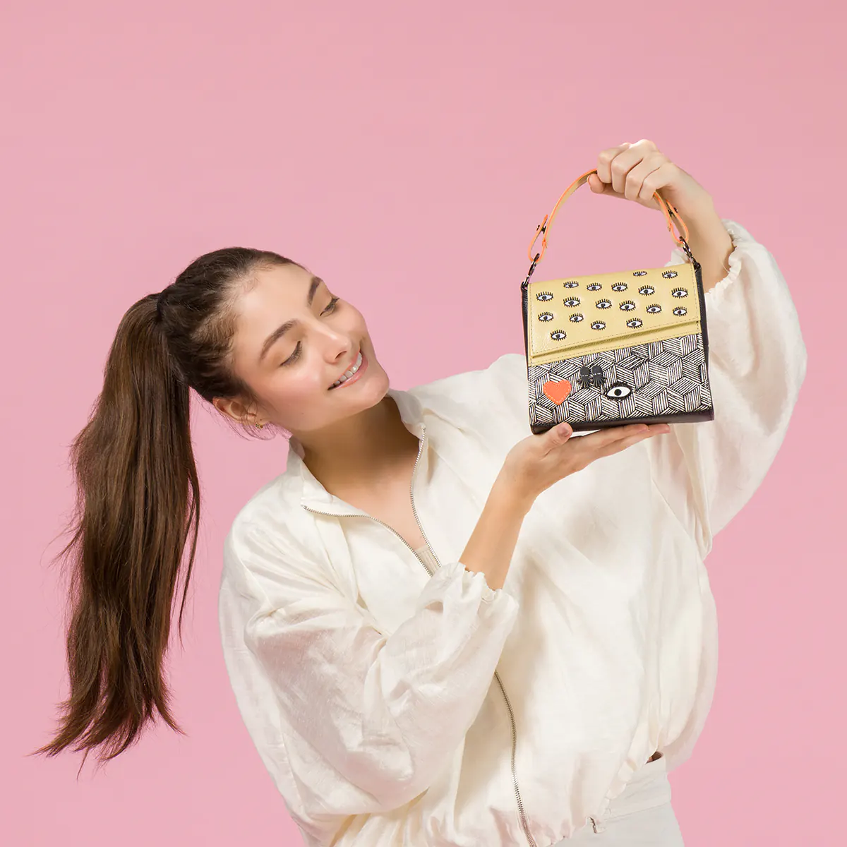 A smiling model in a white blouse holds a Min and Mon handbag featuring a yellow textured flap with eye motifs, a black-and-white geometric pattern, and a red heart patch. Pastel pink background.