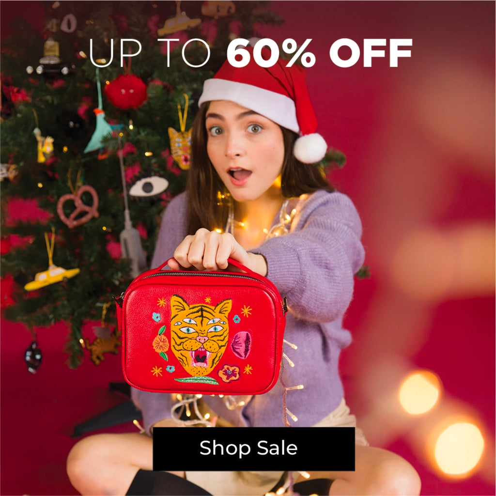 Model holding a red tiger-embroidered Min & Mon bag in front of a Christmas tree, highlighting up to 60% off Sale. Holiday scene with Min & Mon red handbag, festive lights, and “Up to 60% Off” Sale message.