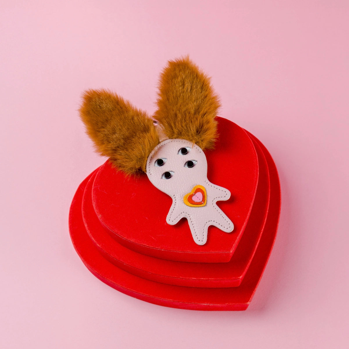 Red heart-shaped box with a whimsical Min & Mon leather charm featuring furry ears and a tiny heart detail on a pink background.