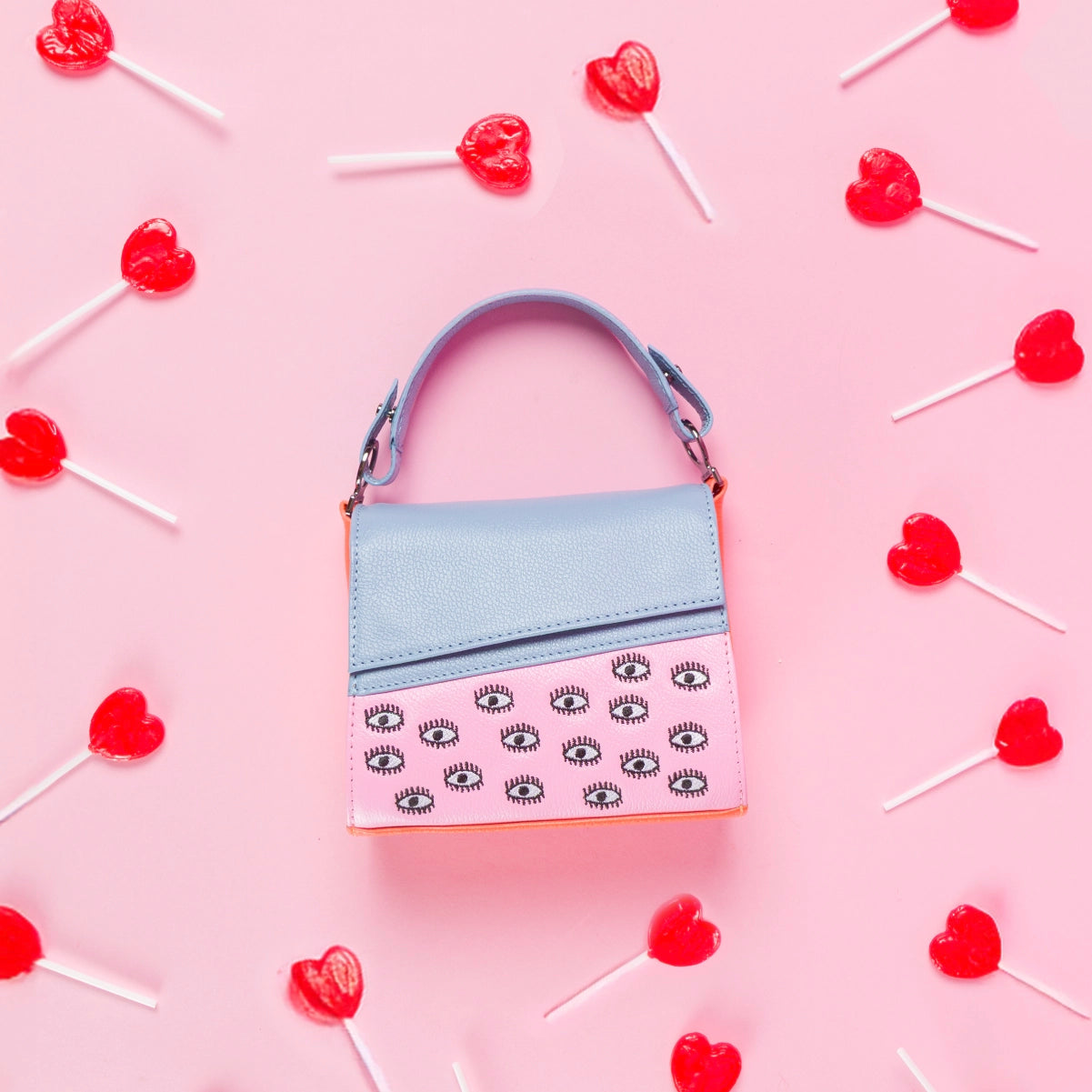 Mini shoulder bag in pastel blue and pink with embroidered eyes, styled on a pink background with red heart-shaped lollipops scattered around.