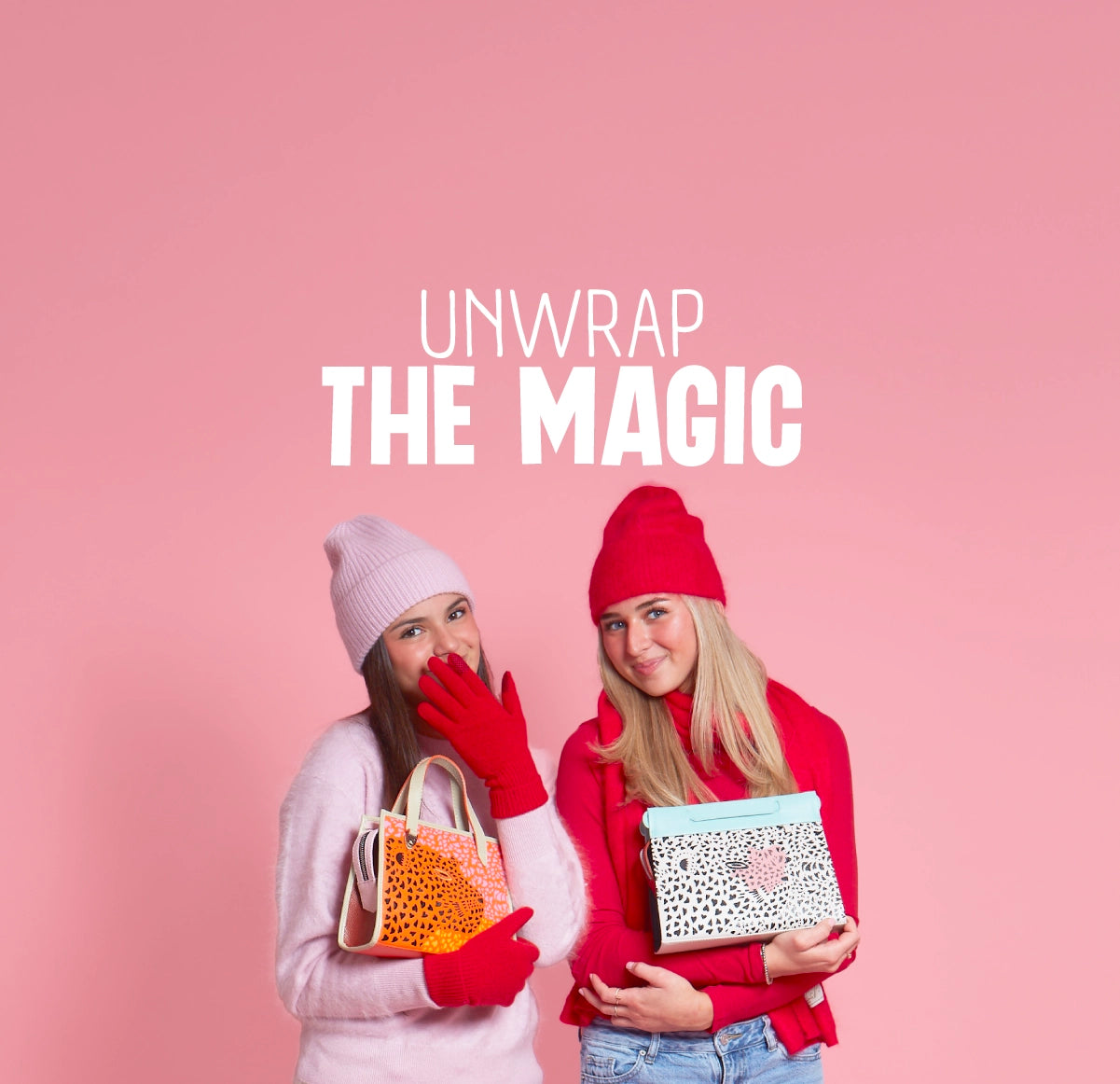 Two smiling young women wearing winter hats and gloves holding colorful Min & Mon bags against a pink holiday background. The banner reads “Unwrap the Magic.”