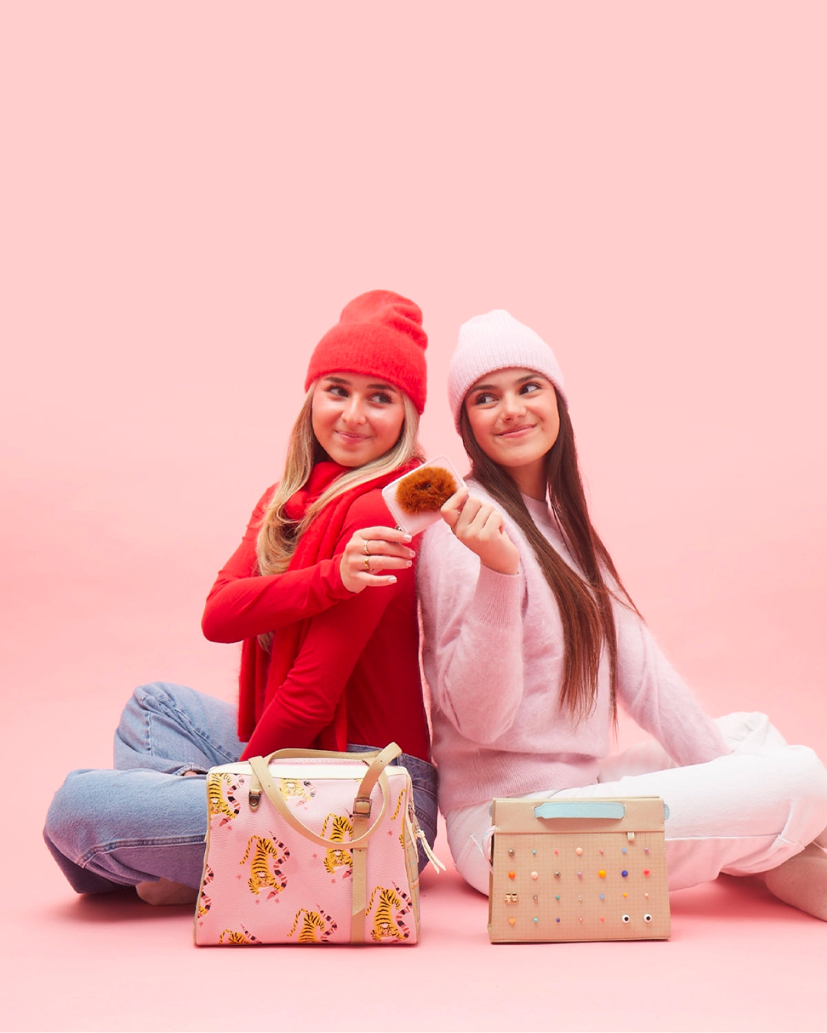 Two women in winter outfits pose back to back on a pink background, showcasing pastel Min & Mon bags and colorful accessories.