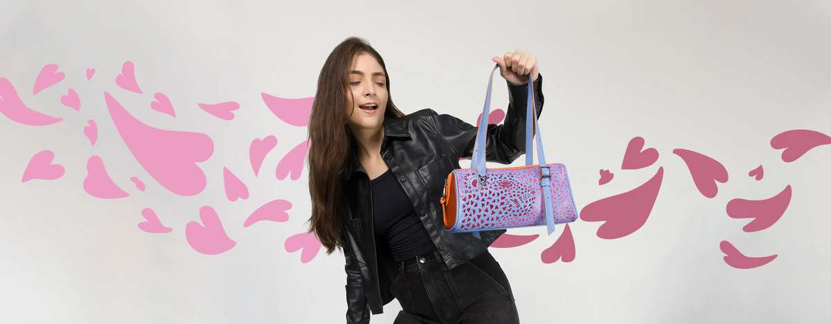 Excited model in a black leather jacket holding the Min and Mon Esther barrel bag in light blue leather with orange accents and a laser-cut heart design. Pink hearts background. New Arrivals banner.