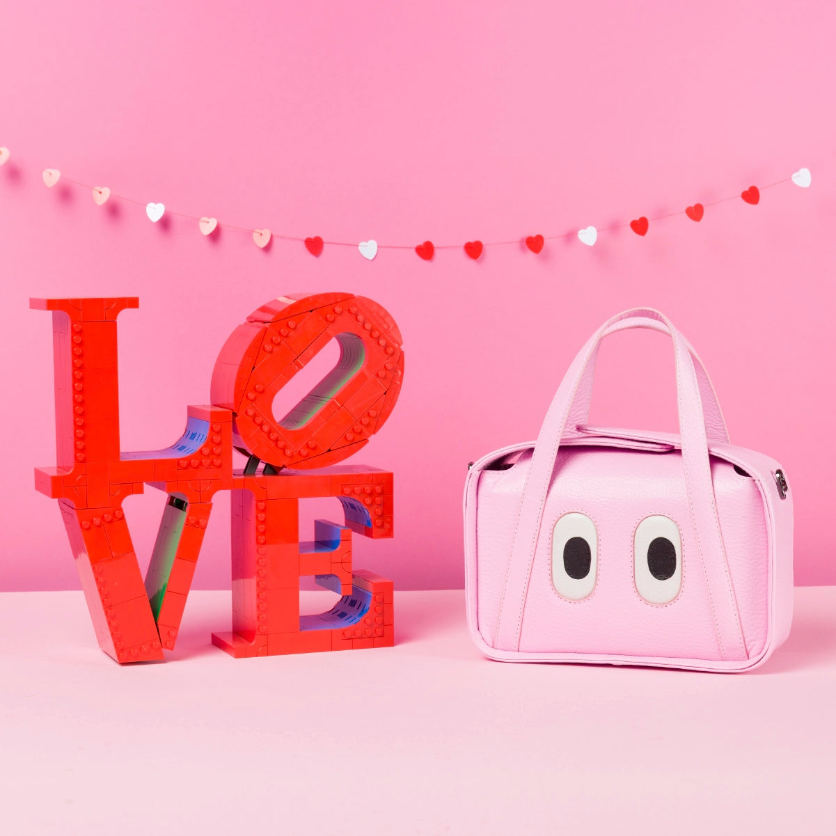 Pink leather handbag with eye appliqué, styled next to red LOVE letters on a pink background with heart garland.
