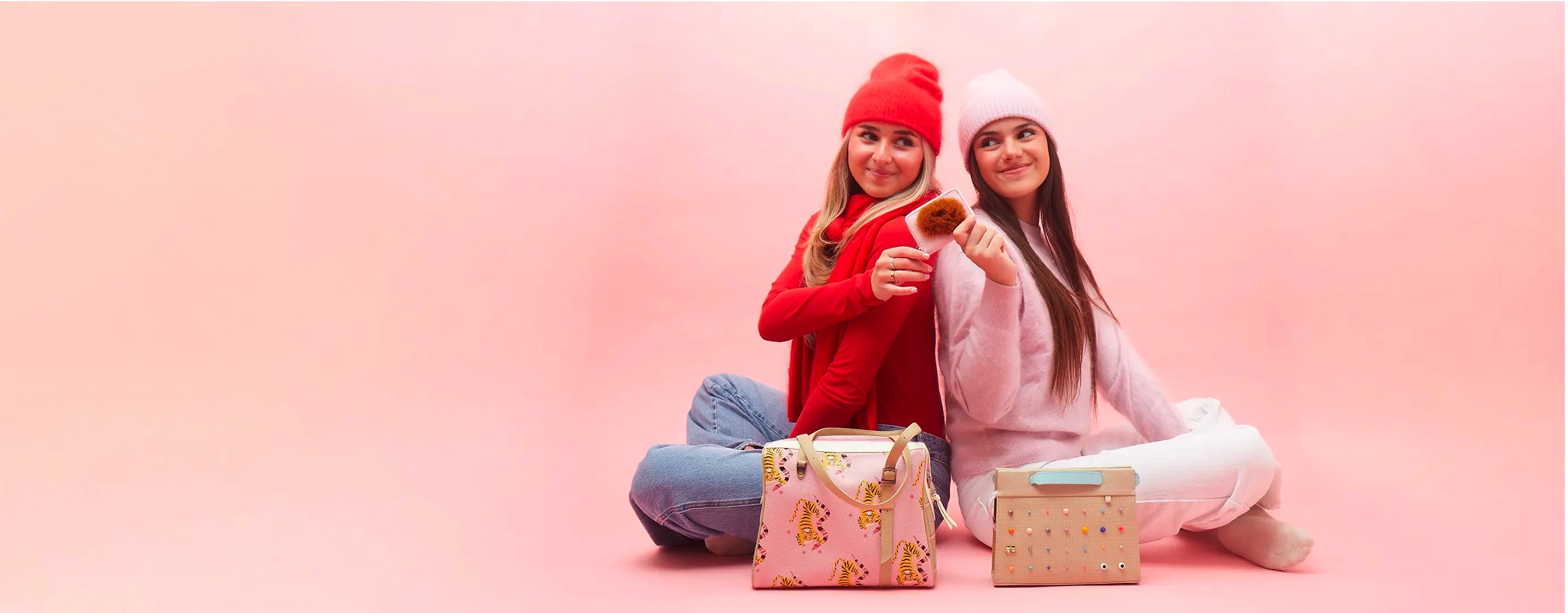 Two women sitting against a pink background wearing winter hats, holding small accessories, with pink Min & Mon bags featuring playful prints.