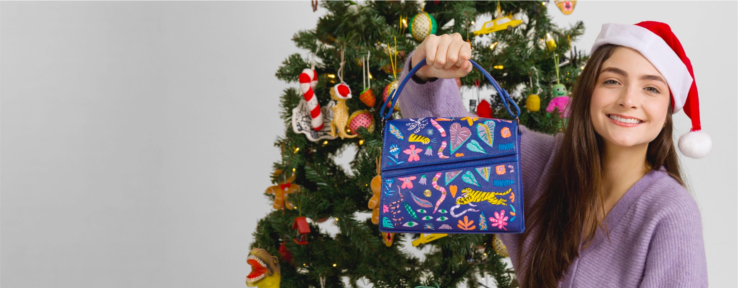 Young woman wearing a Santa hat holding a blue embroidered Min & Mon handbag in front of a decorated Christmas tree.