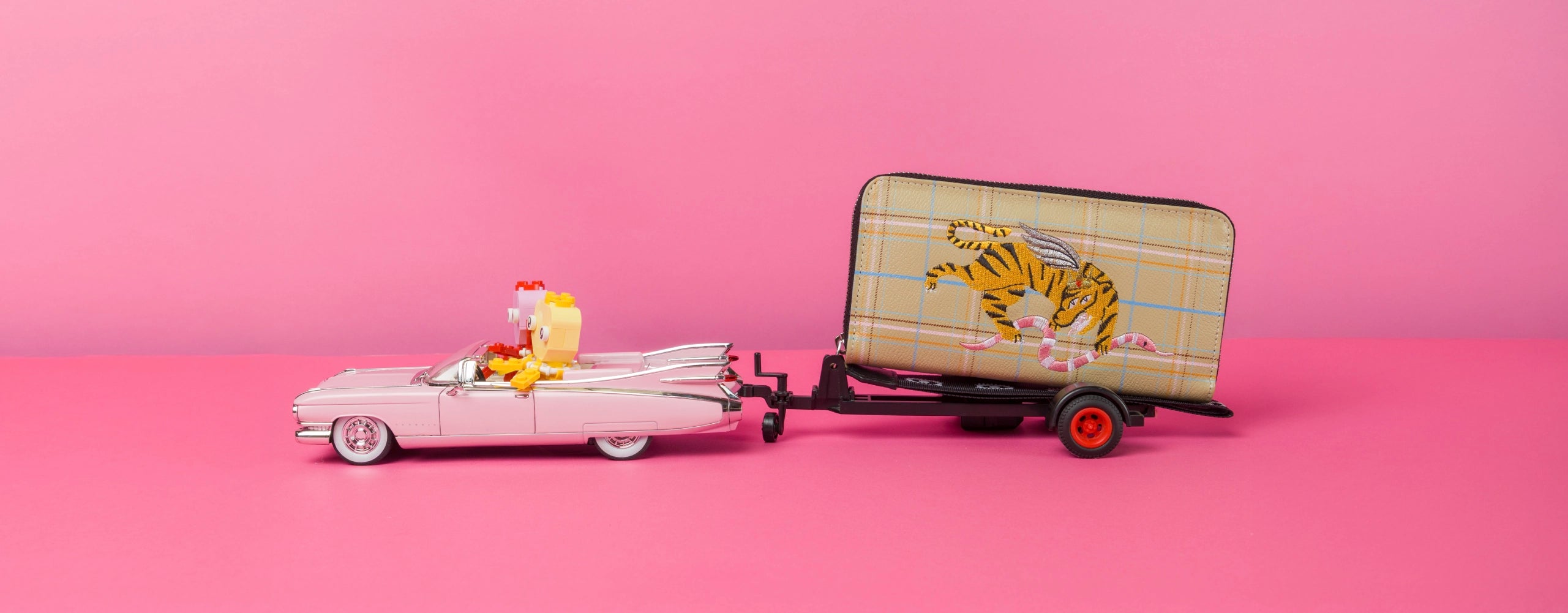 Playful scene of a vintage pink toy car pulling a Min & Mon embroidered tiger wallet on a small trailer over a pink background.