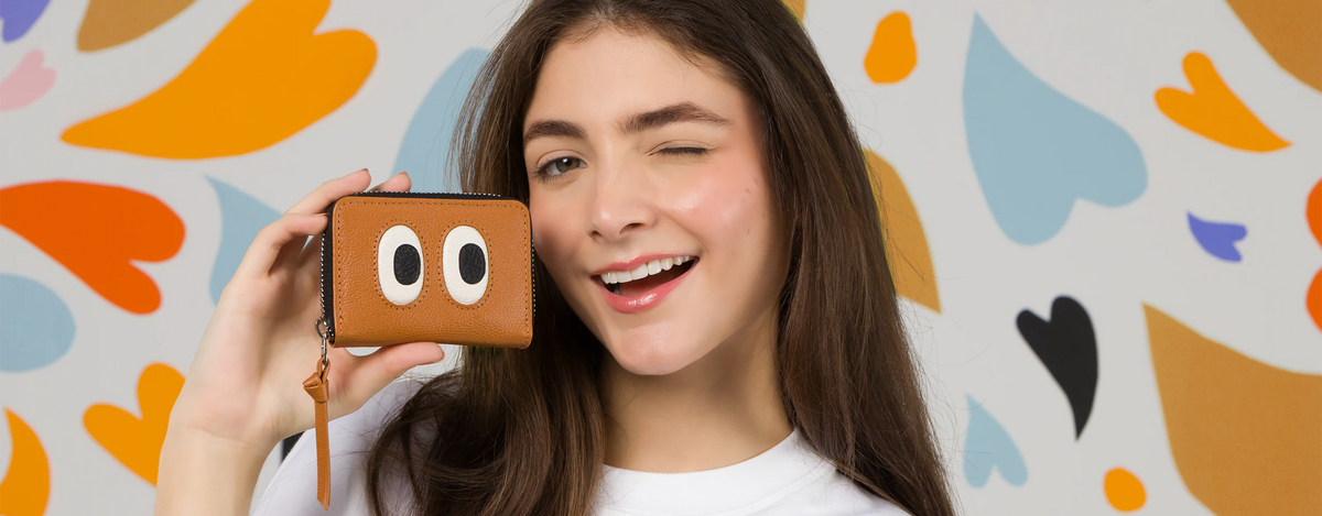  Model winking while holding the Min and Mon Frodo wallet in brown leather, featuring a large eyes design from the Look Up collection. Background with colorful hearts. Wallets section banner.