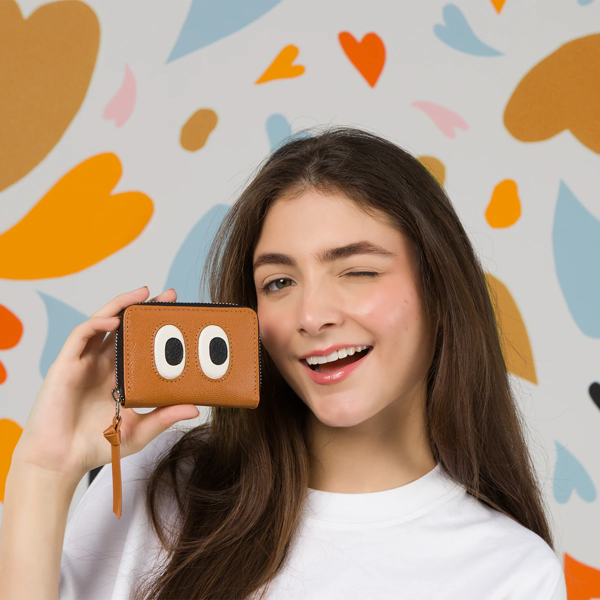  Model winking while holding the Min and Mon Frodo wallet in brown leather, featuring a large eyes design from the Look Up collection. Background with colorful hearts. Wallets section banner.