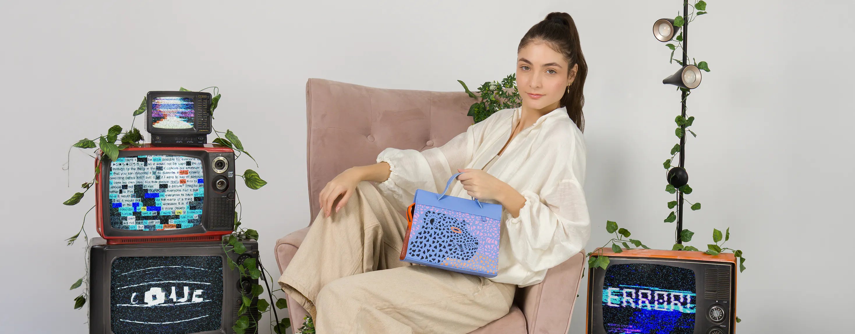 Model sitting in a pink armchair holding the Min and Mon Vali handbag featuring a laser-cut jaguar design for the Wild Heart collection. Retro set with vintage televisions and climbing plants.