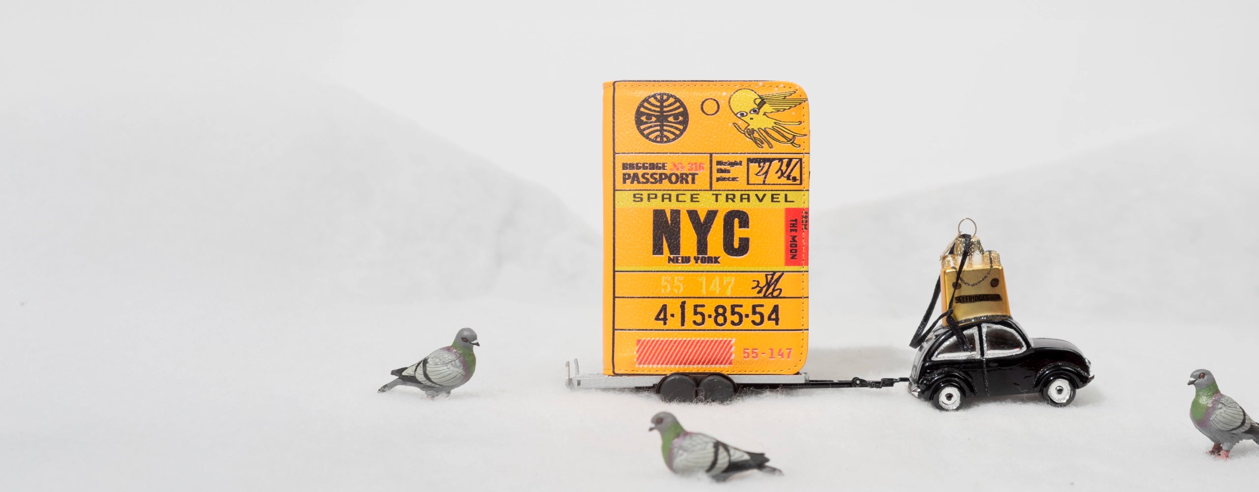 Yellow passport-style wallet with NYC design on a small cart, surrounded by tiny pigeon figures in a winter scene.