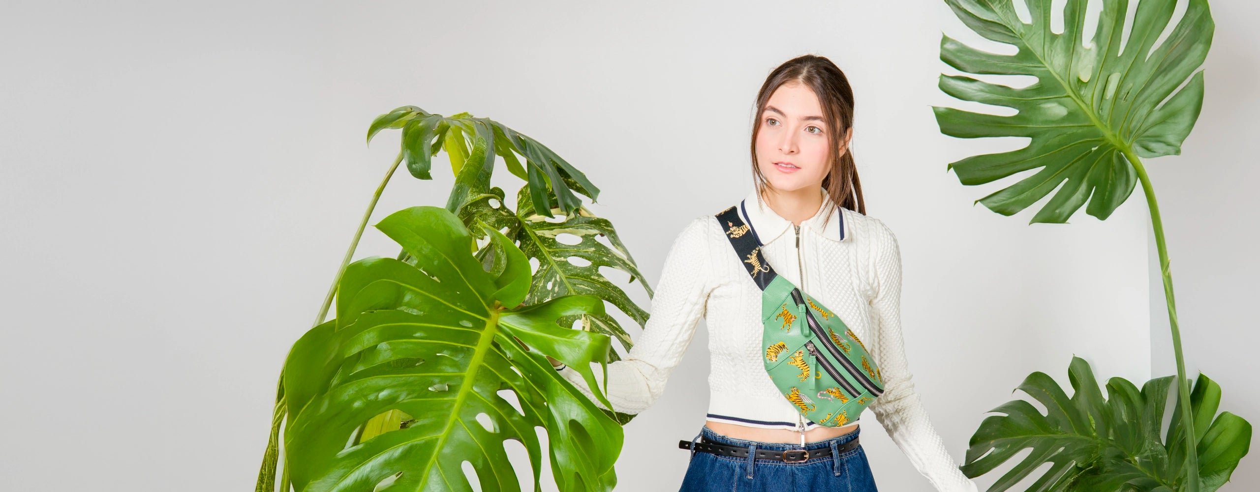 A young woman wearing a textured white zip-up top and high-waisted denim jeans stands in a bright studio surrounded by oversized green monstera leaves. She wears a green crossbody belt bag with embroidered yellow tigers and a thick black strap also decorated with tigers.