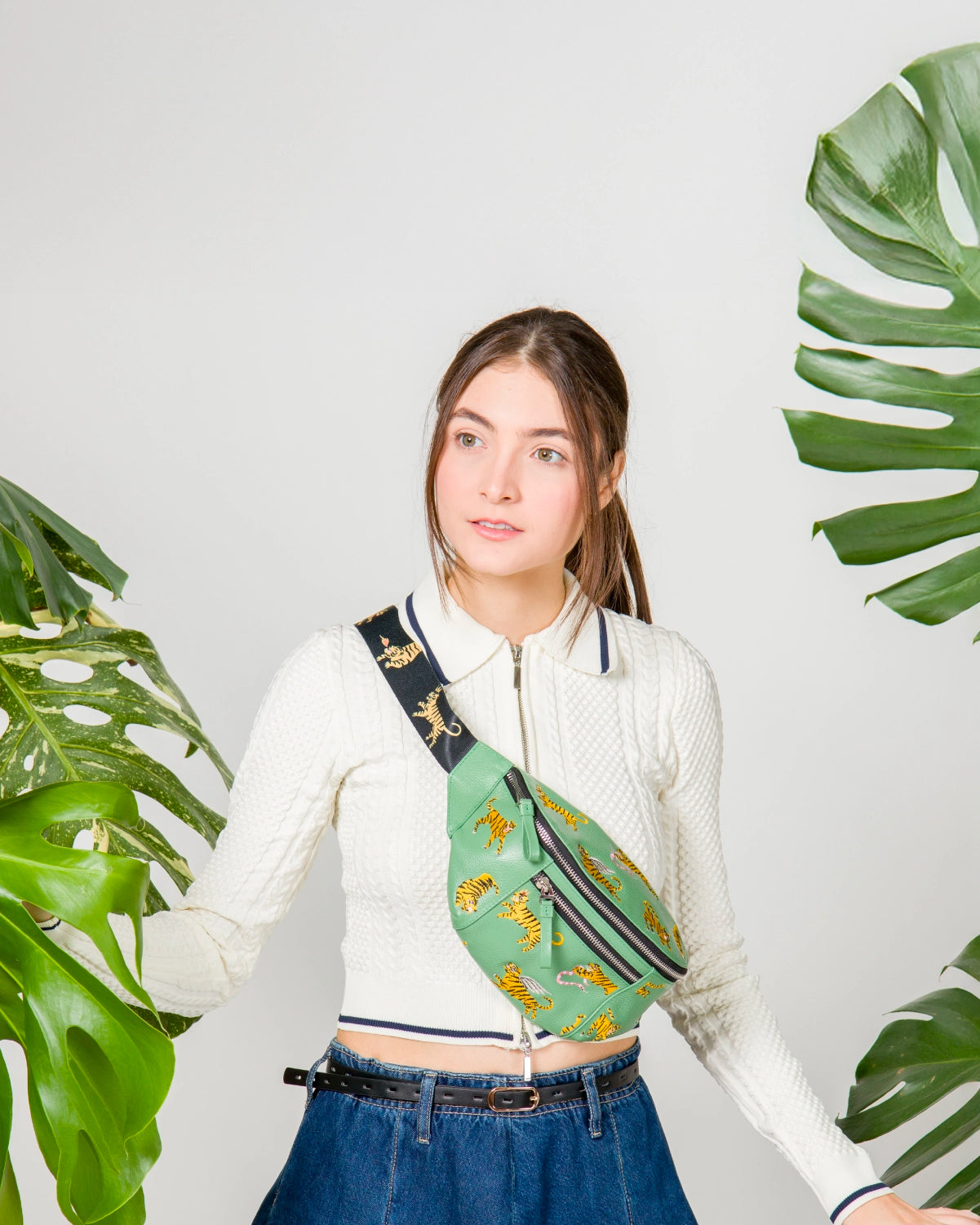 A young woman wearing a textured white zip-up top and high-waisted denim jeans stands in a bright studio surrounded by oversized green monstera leaves. She wears a green crossbody belt bag with embroidered yellow tigers and a thick black strap also decorated with tigers.