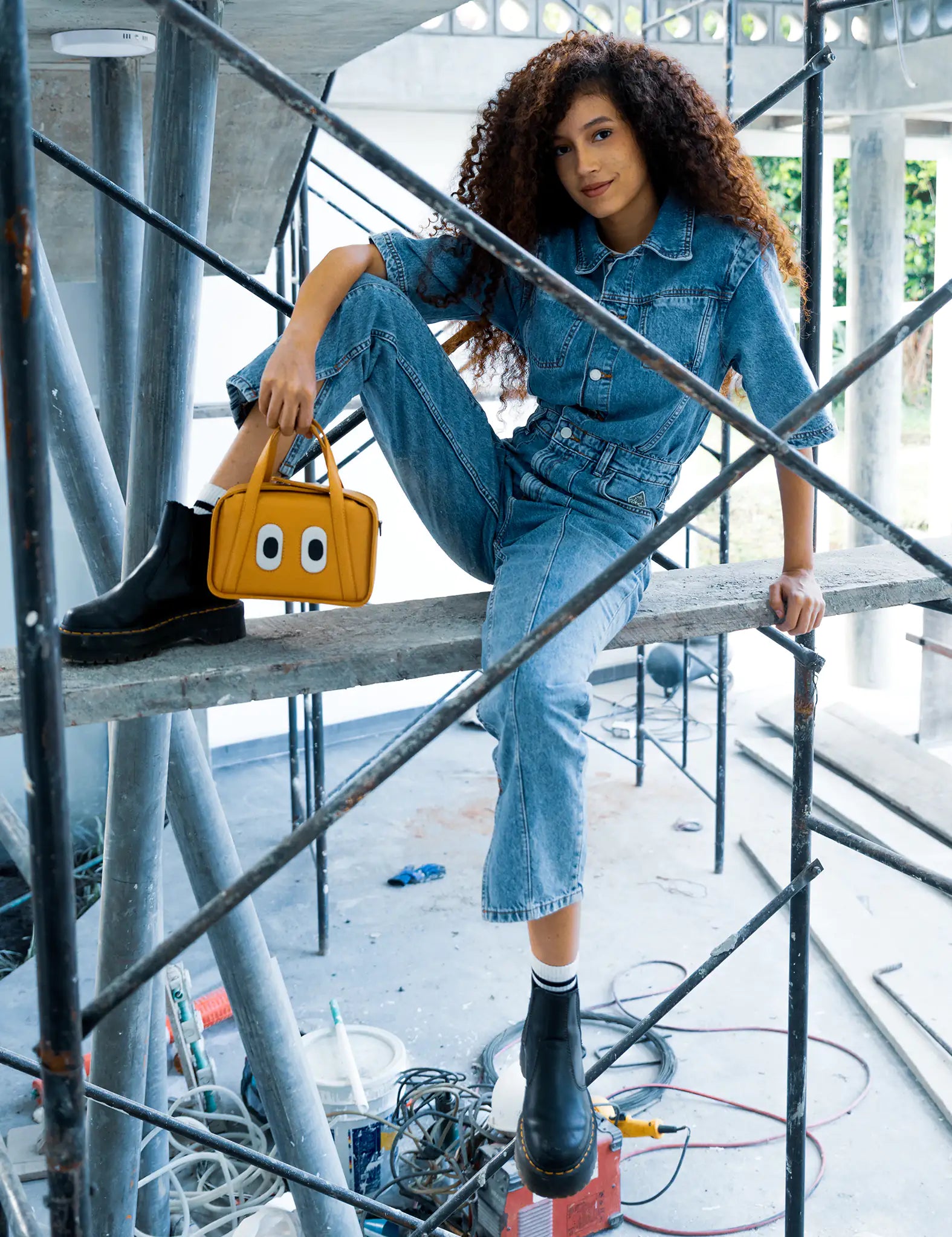 Woman in a denim jumpsuit sits on metal scaffolding, wearing black boots and holding a small mustard-yellow handbag with playful eye details in an industrial setting.