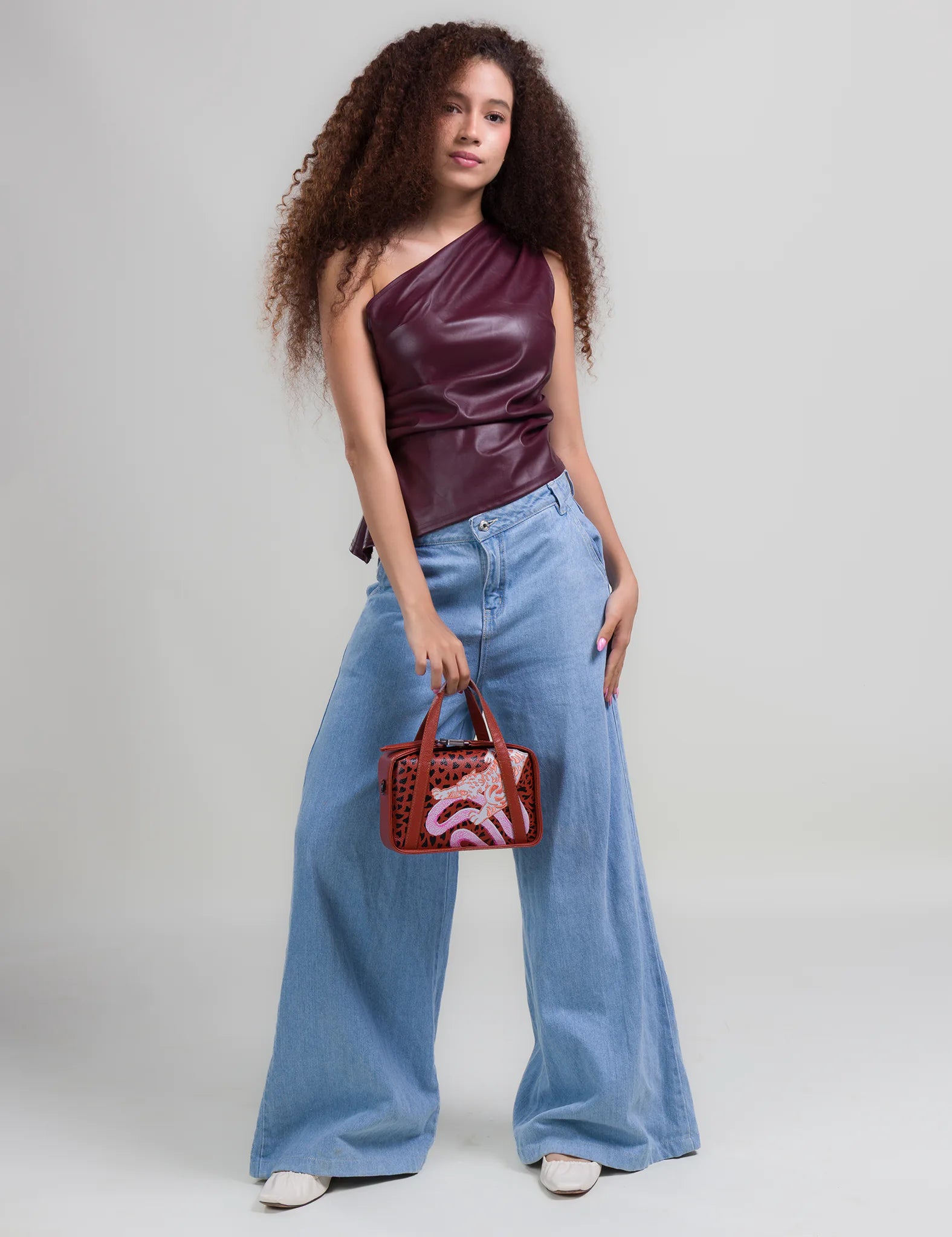 Woman with curly hair wears a burgundy one-shoulder top and wide-leg jeans, holding a small structured handbag with a cutout design and orange and pink tiger & snake motif against a neutral background.