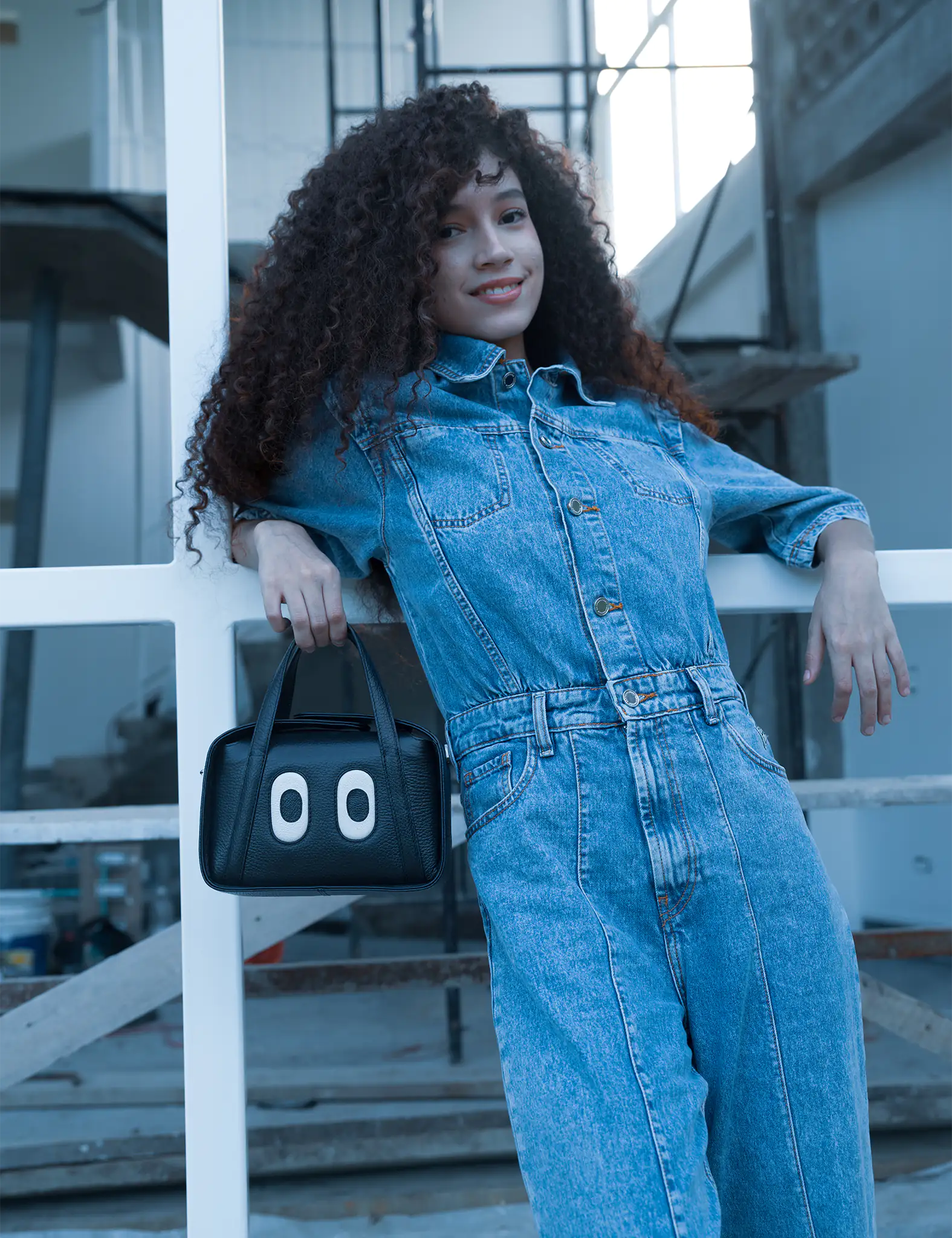 Woman with curly hair in a denim jumpsuit leans against a white railing, smiling while holding a small black handbag with playful eye designs in an industrial setting.