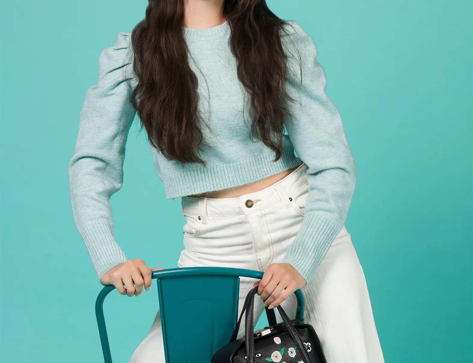 Woman in a light blue sweater and white pants sits on a teal chair, holding a small black handbag with colorful embroidered floral and eye motifs against a matching teal background.