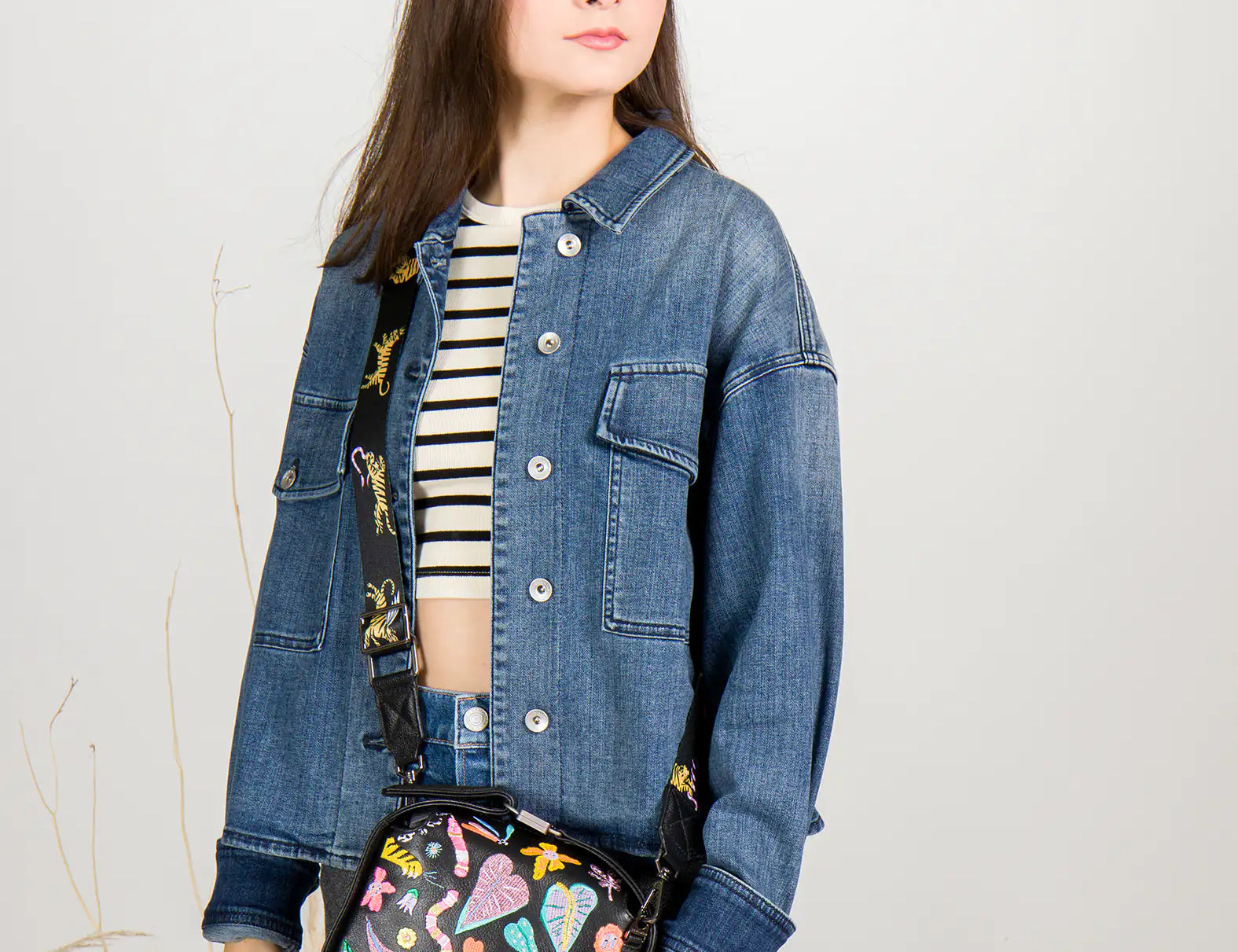 Woman wearing a denim jacket holding a colorful handbag against a plain background