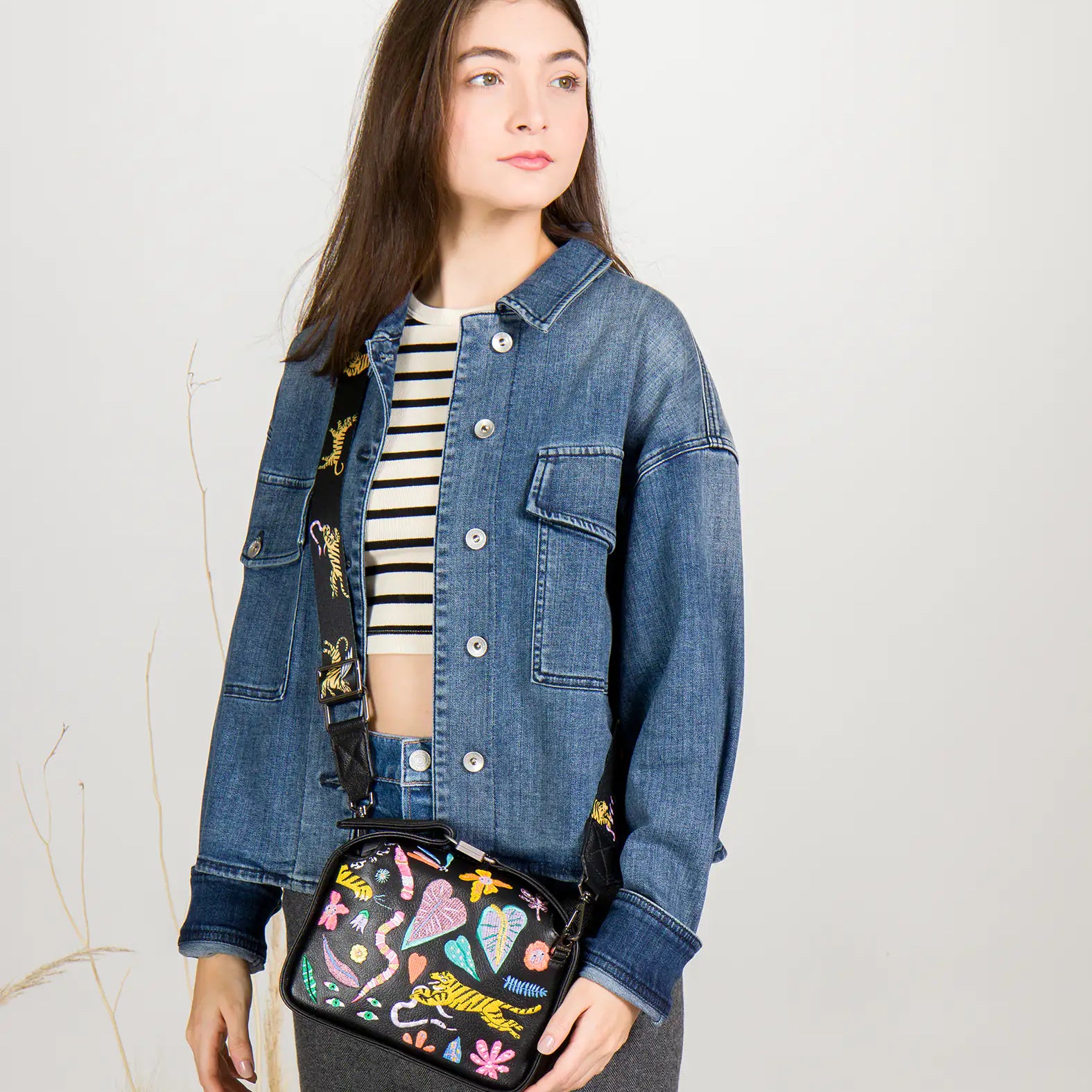 Woman wearing a denim jacket holding a colorful handbag against a plain background