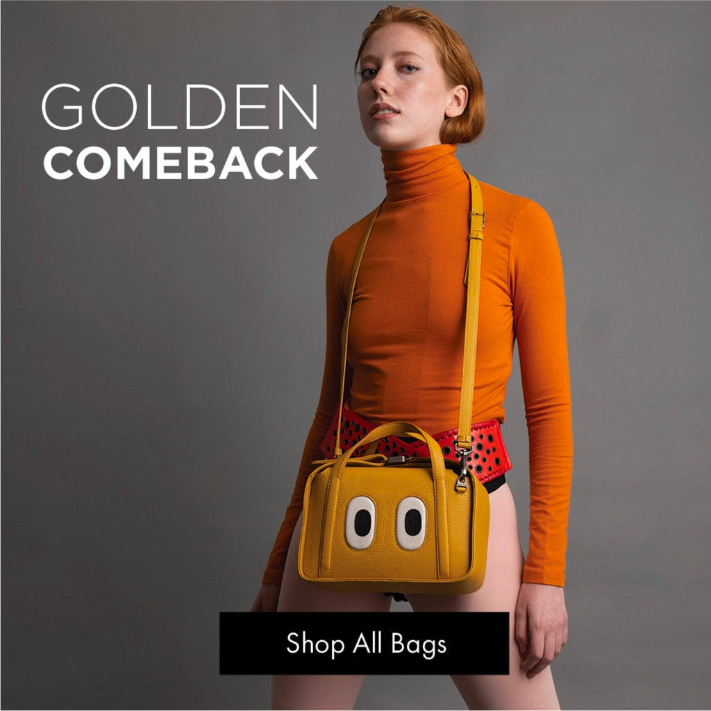 Model wearing an orange turtleneck and holding the yellow Benji Golden handbag by Min & Mon against a gray background.
