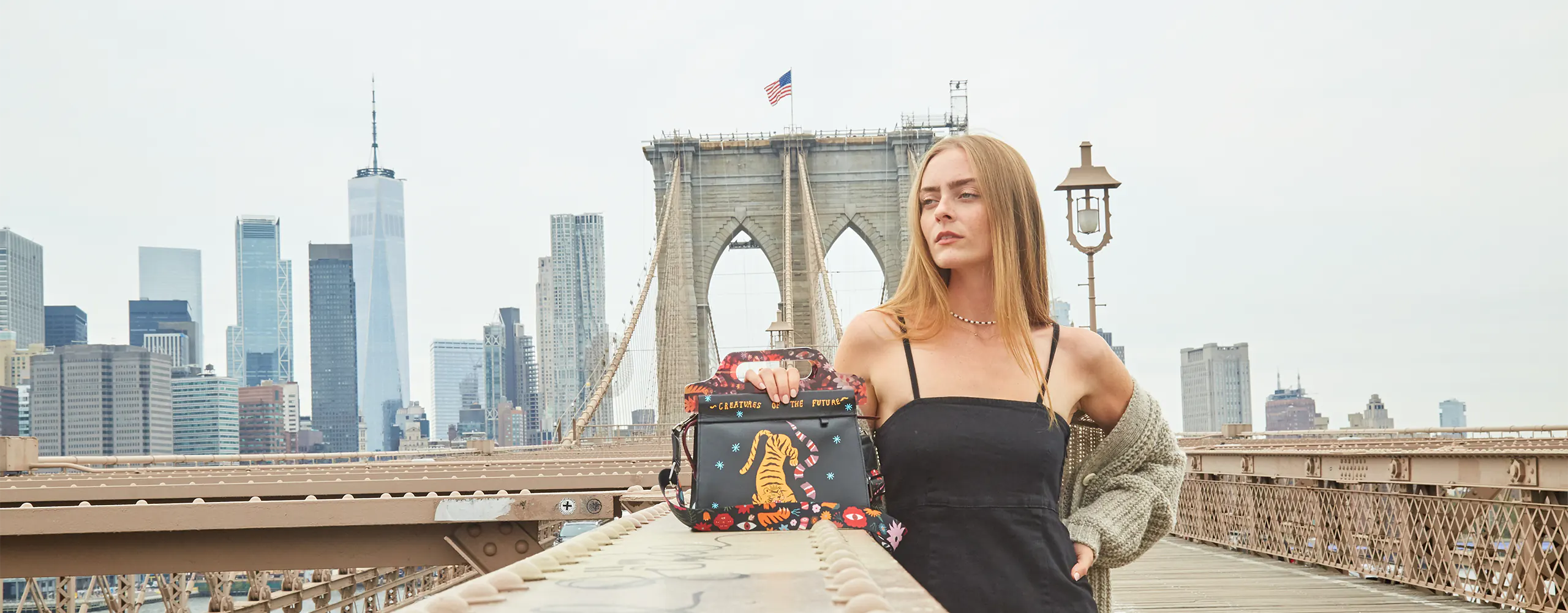 Woman posing on the Brooklyn Bridge with a playful Creatures of the Future handbag and the NYC skyline behind her.