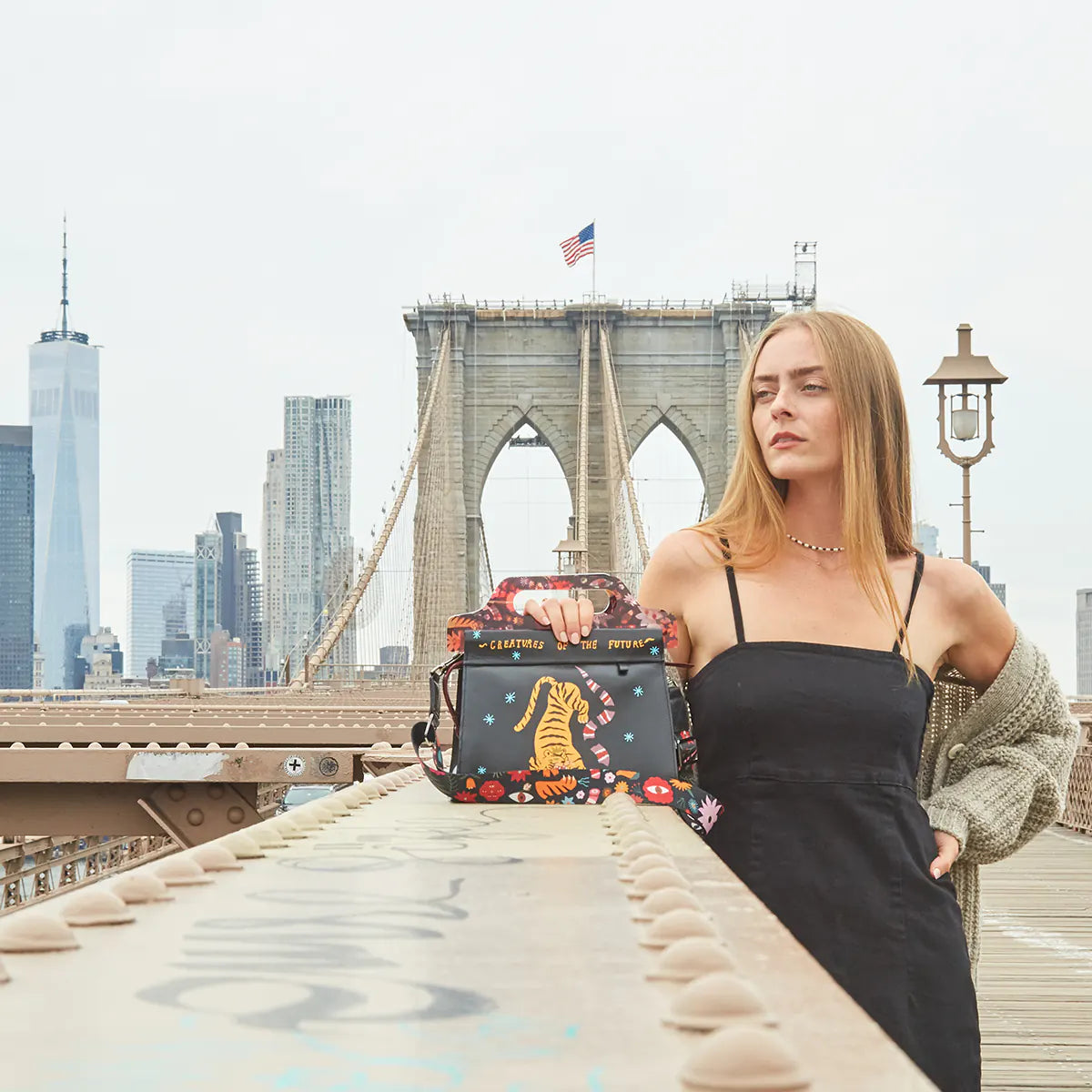 Woman posing on the Brooklyn Bridge with a playful Creatures of the Future handbag and the NYC skyline behind her.